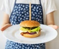 A female chef in an apron shows how she serves a traditional hamburger with cheddar cheese and salad. Close-up