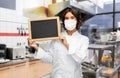 Female chef in mask with chalkboard at kitchen