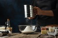 Female chef sifts flour into glass bowl on wooden table with variety of ingredients on dark blue background. Backstage of Royalty Free Stock Photo