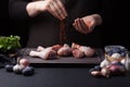A female chef sprinkles fresh raw chicken drumsticks on a dark background. Nearby lie the ingredients for cooking
