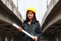 Female civil engineer with yellow helmet, standing with project drafts while in hand on parallel expressway background