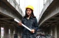 Female civil engineer with yellow helmet, standing with project drafts while in hand on parallel expressway background