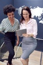 Female Colleagues With Digital Tablet Meeting On Stairs Of Office With World Map In Background