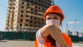 Female construction worker in overalls and medical mask coughing in elbow on background of house under construction