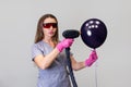 Female cosmetologist holds black balloon and directs handle of laser device into it. Gray background. Concept of laser