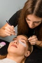 Female cosmetologist with tweezers attaches artificial eyelashes to female client at salon.