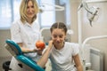Female dentist gives apple for smiling girl in modern dental room.