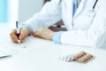 Female doctor filling up prescription form while sitting at the desk in hospital. Close-up of medical pills, lying at