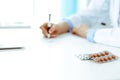 Female doctor filling up prescription form while sitting at the desk in hospital. Close-up of medical pills, lying at