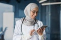 Female doctor in hijab works in modern clinic office, Muslim female doctor uses tablet computer, nurse in medical white Royalty Free Stock Photo