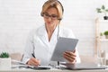 Female doctor makes notes in notebook, holds tablet in hand in interior of office