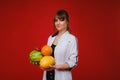 A female doctor nurse in a white coat with fruit in her hands poses on a red background, melon, watermelon, strawberry and