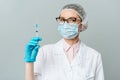 Female doctor or scientist in white medical gown, blue gloves, green cap and mask holds a syringe in hands on white