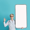 Female doctor showing big smartphone with blank screen and gesturing OK sign, standing over blue background, mockup Royalty Free Stock Photo