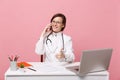 Female doctor sit at desk work on computer with medical document hold cellphone in hospital isolated on pastel pink Royalty Free Stock Photo