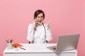 Female doctor sit at desk work on computer with medical document hold cellphone in hospital isolated on pastel pink Royalty Free Stock Photo