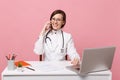 Female doctor sit at desk work on computer with medical document hold cellphone in hospital isolated on pastel pink Royalty Free Stock Photo