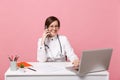 Female doctor sit at desk work on computer with medical document hold cellphone in hospital isolated on pastel pink Royalty Free Stock Photo