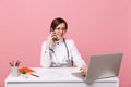 Female doctor sit at desk work on computer with medical document hold cellphone in hospital isolated on pastel pink Royalty Free Stock Photo