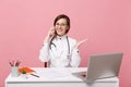 Female doctor sit at desk work on computer with medical document hold cellphone in hospital isolated on pastel pink Royalty Free Stock Photo