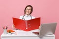 Female doctor sit at desk work on computer with medical document hold folder in hospital isolated on pastel pink wall Royalty Free Stock Photo