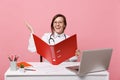 Female doctor sit at desk work on computer with medical document hold folder in hospital isolated on pastel pink wall Royalty Free Stock Photo