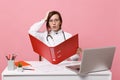 Female doctor sit at desk work on computer with medical document hold folder in hospital isolated on pastel pink wall Royalty Free Stock Photo