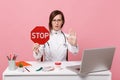 Female doctor sit at desk work on computer with medical document hold stop in hospital isolated on pastel pink wall