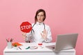 Female doctor sit at desk work on computer with medical document hold stop in hospital isolated on pastel pink wall