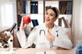 Female doctor sitting at desk in office with microscope and stethoscope. Woman is holding red apple. Royalty Free Stock Photo