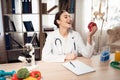 Female doctor sitting at desk in office with microscope and stethoscope. Woman is holding red apple. Royalty Free Stock Photo