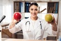 Female doctor sitting at desk in office with microscope and stethoscope. Woman is holding yellow and red apples. Royalty Free Stock Photo