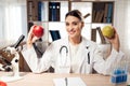 Female doctor sitting at desk in office with microscope and stethoscope. Woman is holding yellow and red apples. Royalty Free Stock Photo