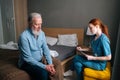 Female doctor talking to senior adult male patient sitting on bed at home and checking health writing symptoms in Royalty Free Stock Photo