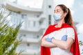 A female doctor wearing a surgical mask and rubber gloves poses with her arms crossed and a super hero's cape on her back