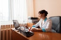 A female doctor in a white bathrobe works behind a laptop near a window in a hospital. Responsible work.