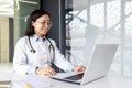 Female doctor working at laptop while sitting at desk and smiling in hospital office. Asian medic woman providing online Royalty Free Stock Photo