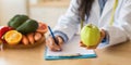 Female doctor writing diet plan and holding green apple, sitting at desk with fresh fruits, cropped, panorama, closeup