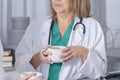 a female doctor and a young nurse keep cups of coffee in hands and gossip with each other during coffee break in the staff room