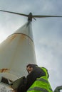 Female engineer with computer in hand and yellow reflective jacket preparing for technical control under a wind turbine