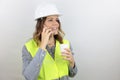 Female engineer wearing reflecting jacket and hardhat smiling happy holding paper cup takeaway speaking on phone. Royalty Free Stock Photo