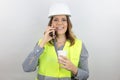 Female engineer wearing reflecting jacket and hardhat smiling happy holding smartphone and paper cup takeaway. Royalty Free Stock Photo