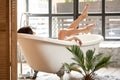 A female enjoy lies in a white bathtub and looks out the street through a large window. Back view shot