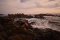 Woman rests on coastal boulders as waves crash nearby, serene dusk engulfs seascape. Female enjoys peaceful ocean sunset