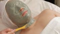The female face is completely covered with a cosmetic mask. The beautician applies a gray mass to the skin of the girl. Middle-