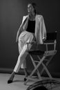 Female Fashion Portrait. Beautiful Woman In White Suit Stands Near The Wooden Director`s Chair.