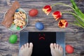 Female feet on digital scales with sad smile surrounded by Easter food Easter cake, chocolate Easter bunny, painted eggs and Royalty Free Stock Photo