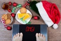 Female feet on digital scales with sign omg! surrounded by Christmas decorations, bottle, glass of alcohol and sweets.