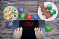 Female feet on digital scales with sign omg! surrounded by Easter food Easter cake, chocolate Easter bunny, painted eggs.