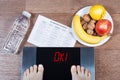 Female feet on digital scales with word ok surrounded by bottle of water, plate with healthy food and workout schedule paper.